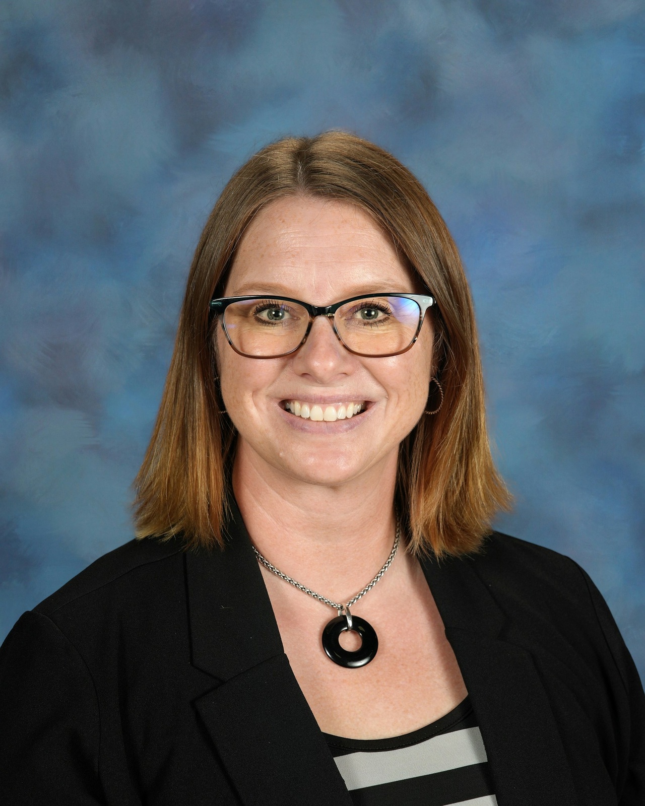 A smiling woman with glasses and shoulder-length brown hair wears a black blazer and necklace, set against a mottled blue background.